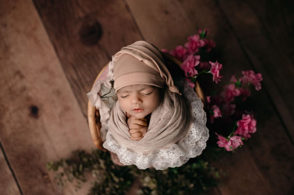 Overhead view of sleeping newborn wrapped in beige swaddle with pink flowers and greenery on wooden floor.