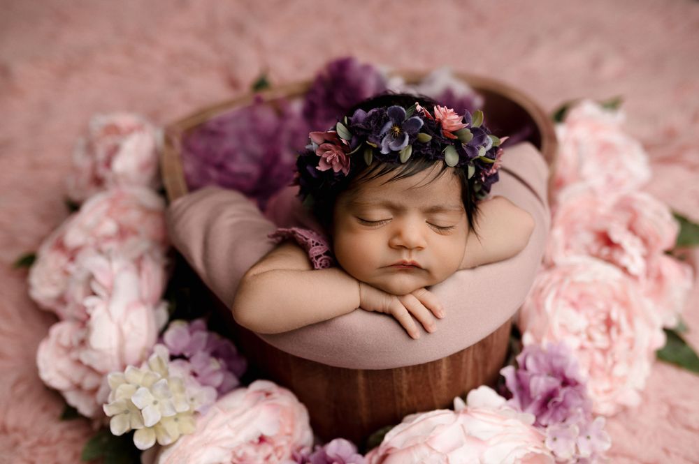 Newborn sleeps peacefully in wooden bowl surrounded by pink and purple flowers and dark floral crown.