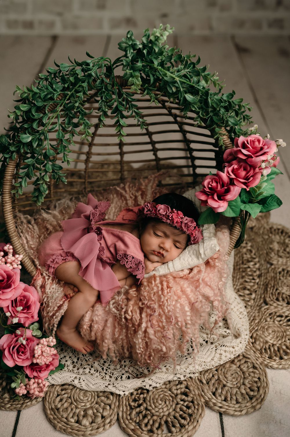 Whimsical newborn photography setup with wicker basket, pink roses, greenery and lace on textured background.
