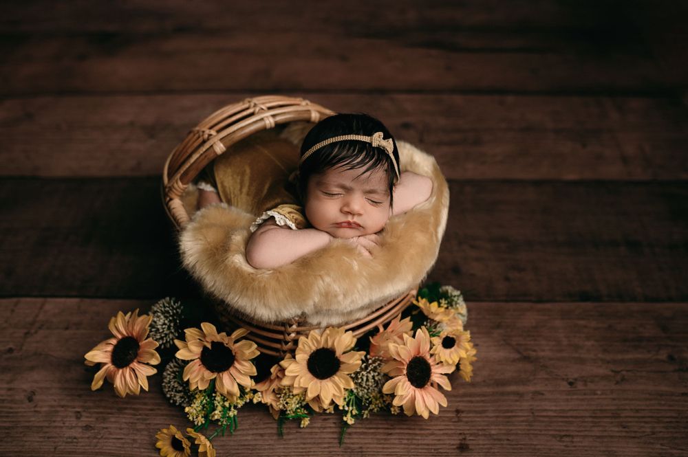 A newborn sleeps peacefully in a brown basket surrounded by bright yellow sunflowers on a wooden floor.