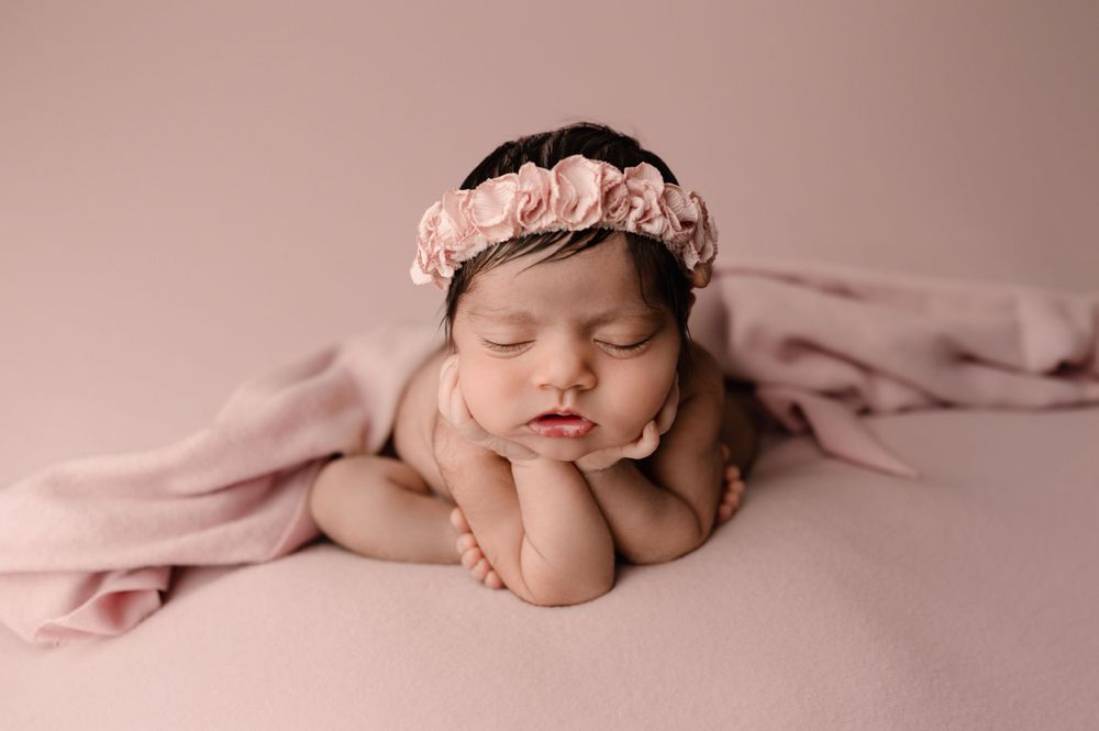 Newborn baby sleeping peacefully on pink blanket wearing delicate flower crown headband.