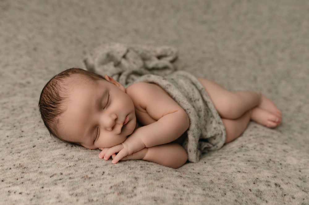 Sleeping newborn baby peacefully resting on beige carpet wrapped in soft grey blanket.