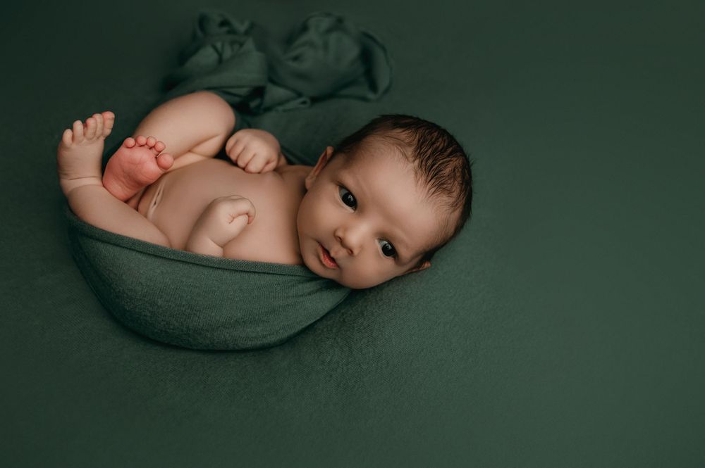 Sweet newborn baby curled up in a green bowl pose during professional photo session on dark green backdrop.