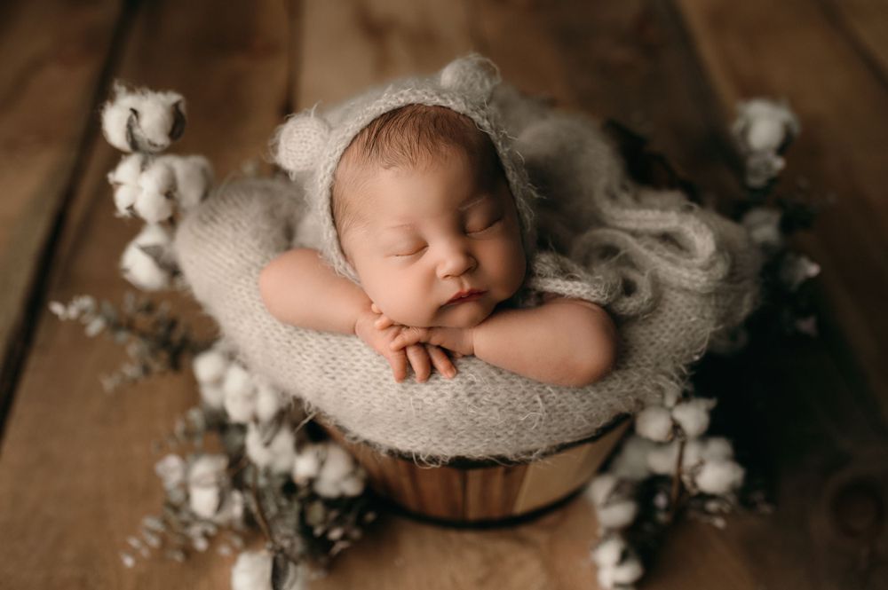 Sleeping newborn peacefully rests in wooden bowl surrounded by soft white fabric and cotton flowers on wooden surface.