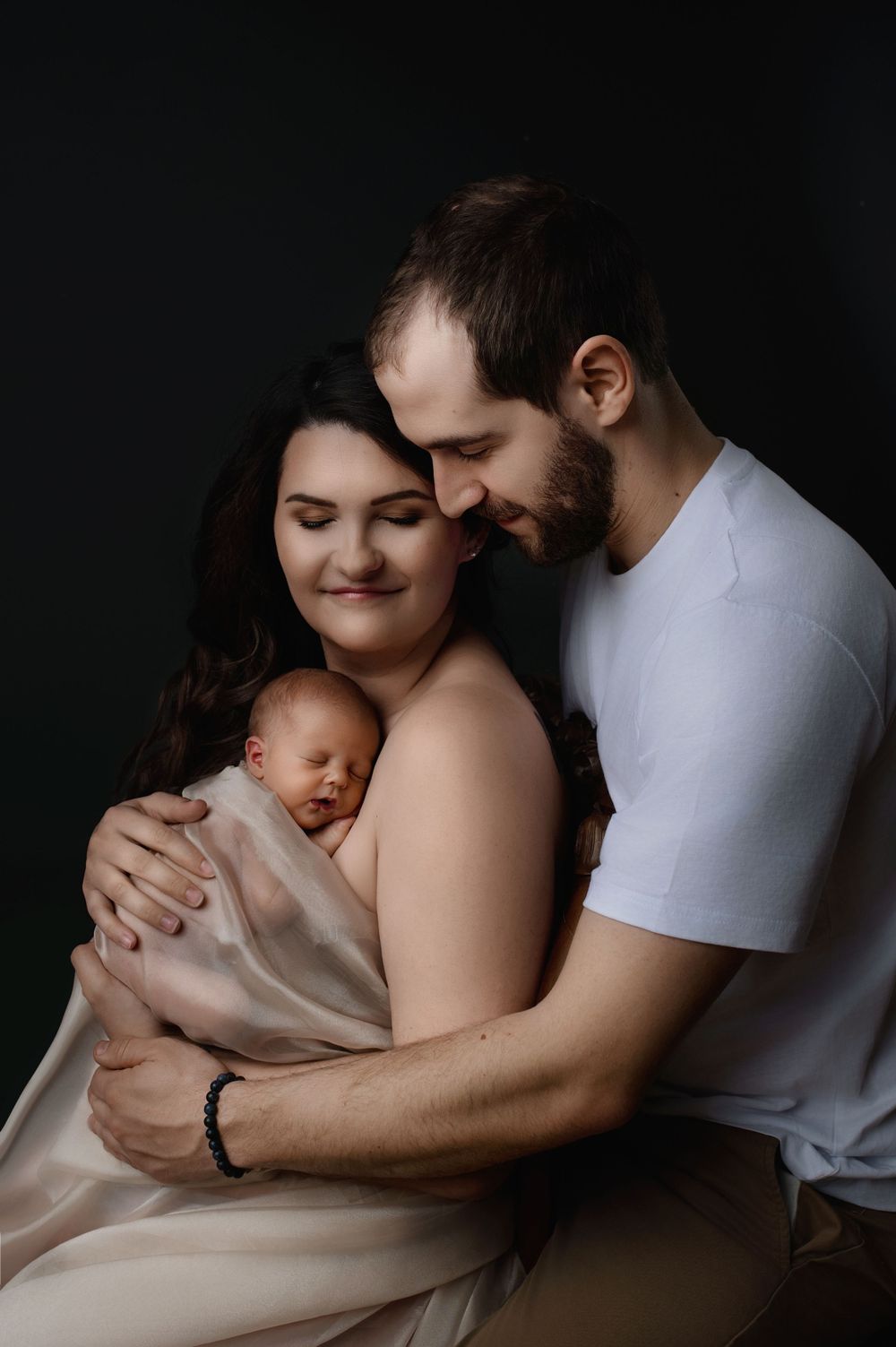 A couple shares an intimate moment with their newborn baby wrapped in beige fabric against a dark background.