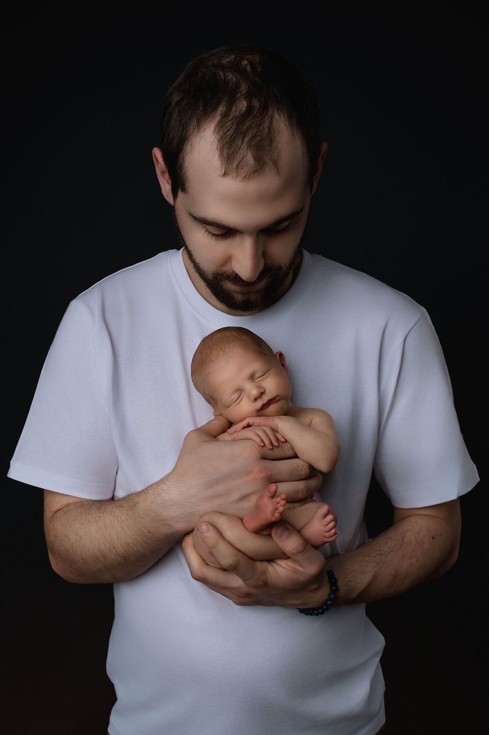 A tender moment shows a man in a white t-shirt gently cradling a sleeping newborn baby against his chest.