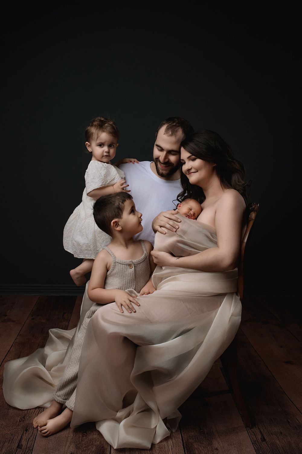 A family of five sits together on a wooden floor in ethereal cream-colored clothing against a dark backdrop.