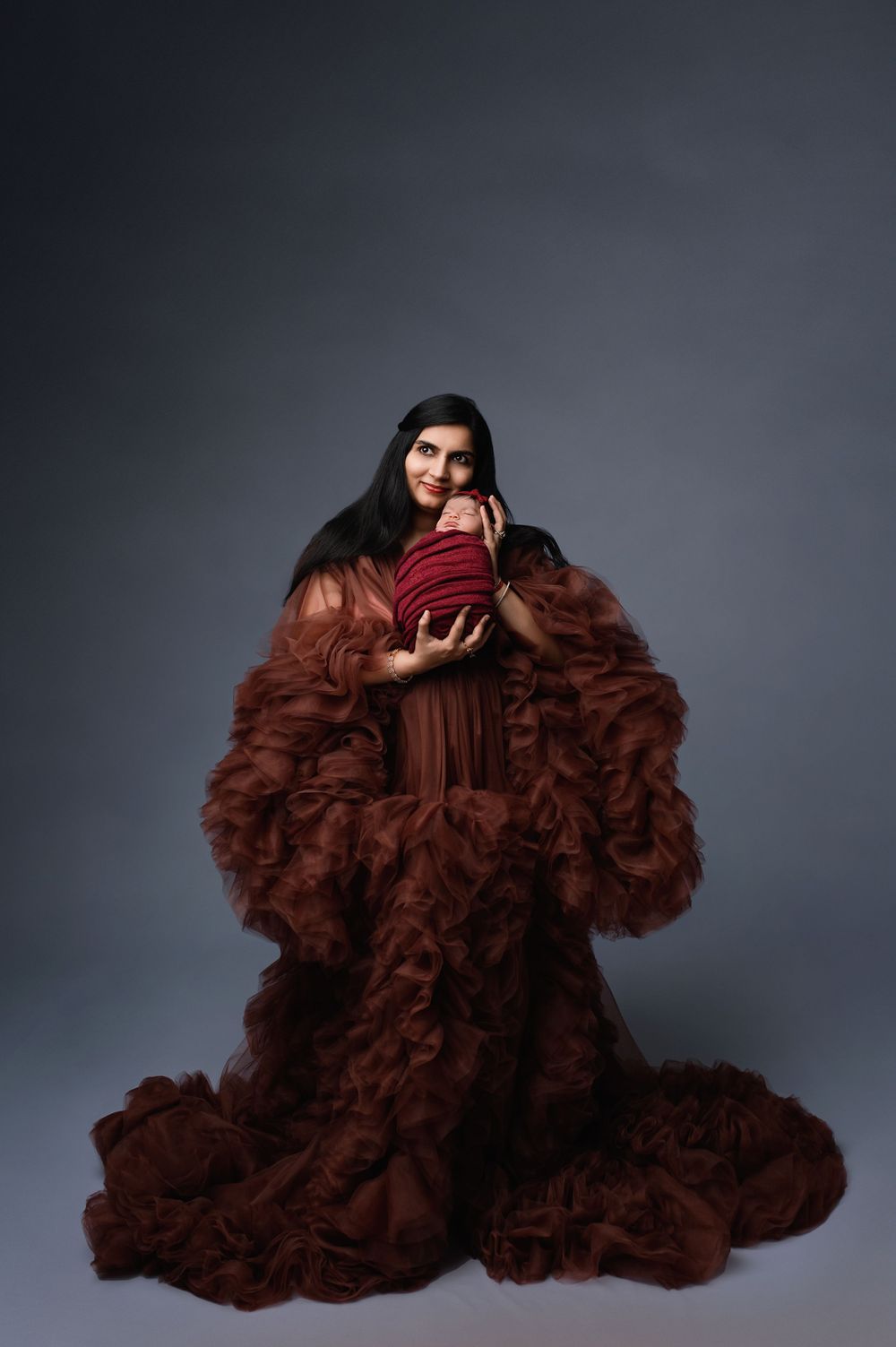 Fashion portrait of someone wearing a dramatic ruffled brown gown and dark red velvet top against a gray background.