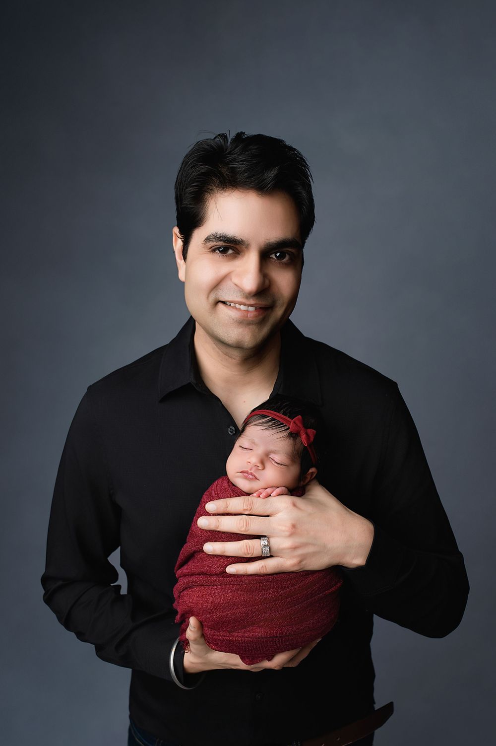 Professional studio portrait features an adult in black holding a sleeping newborn wrapped in burgundy fabric against dark backdrop.