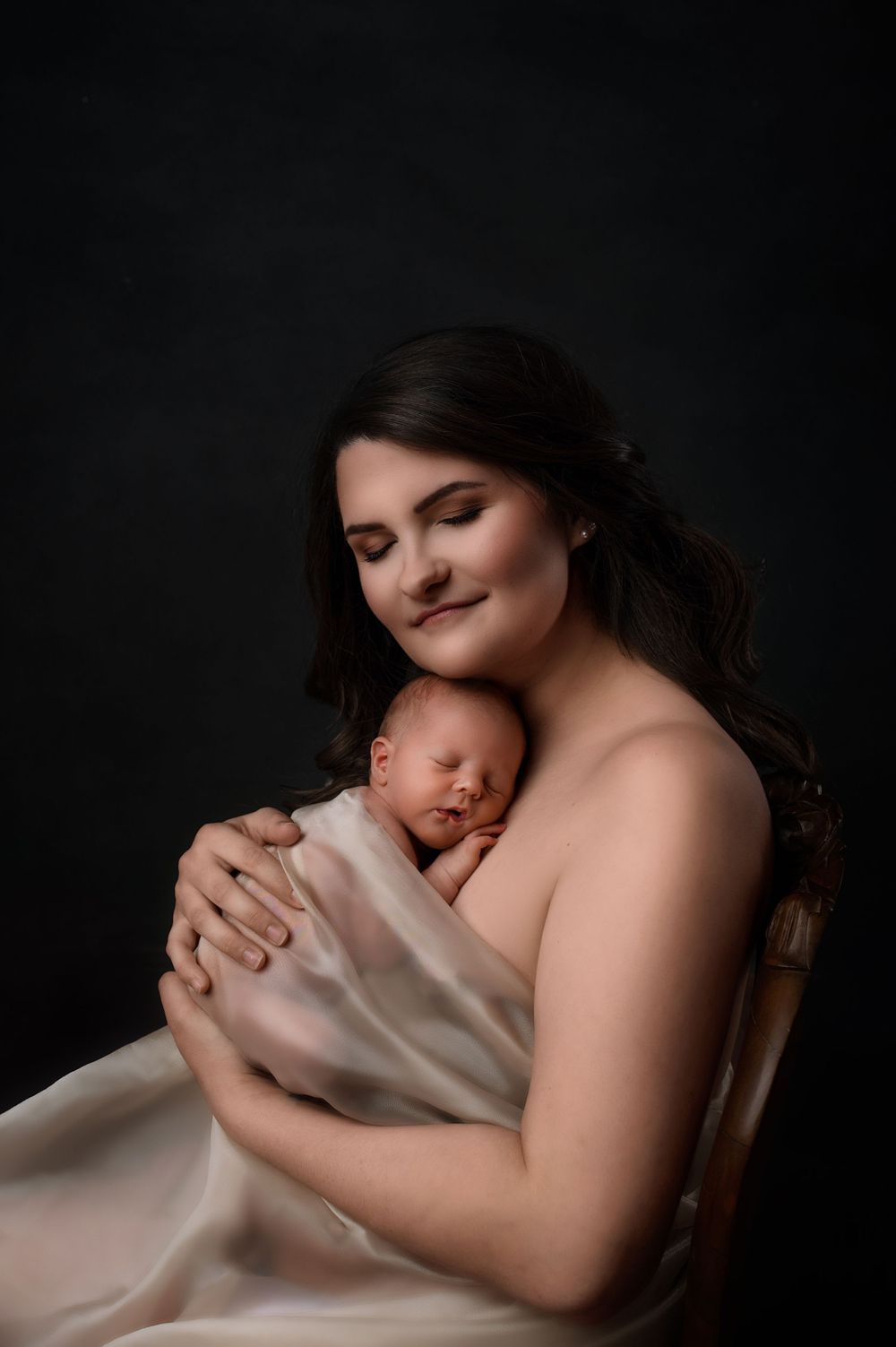 An intimate studio portrait of a tender moment between parent and newborn draped in soft white fabric against a dark background.