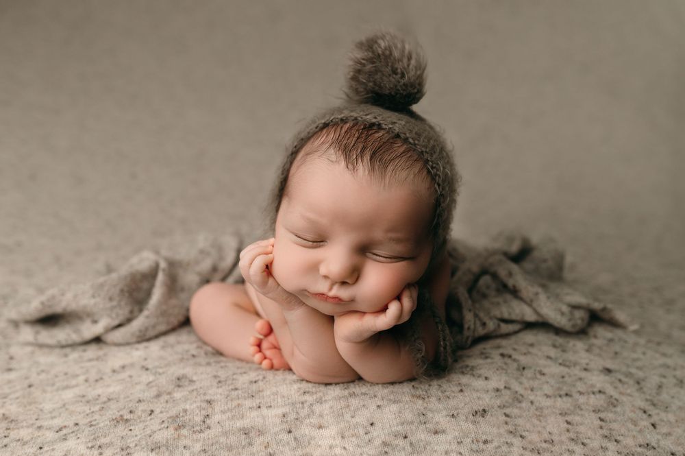 A sleeping newborn rests with tiny fist under chin on soft gray textured backdrop.