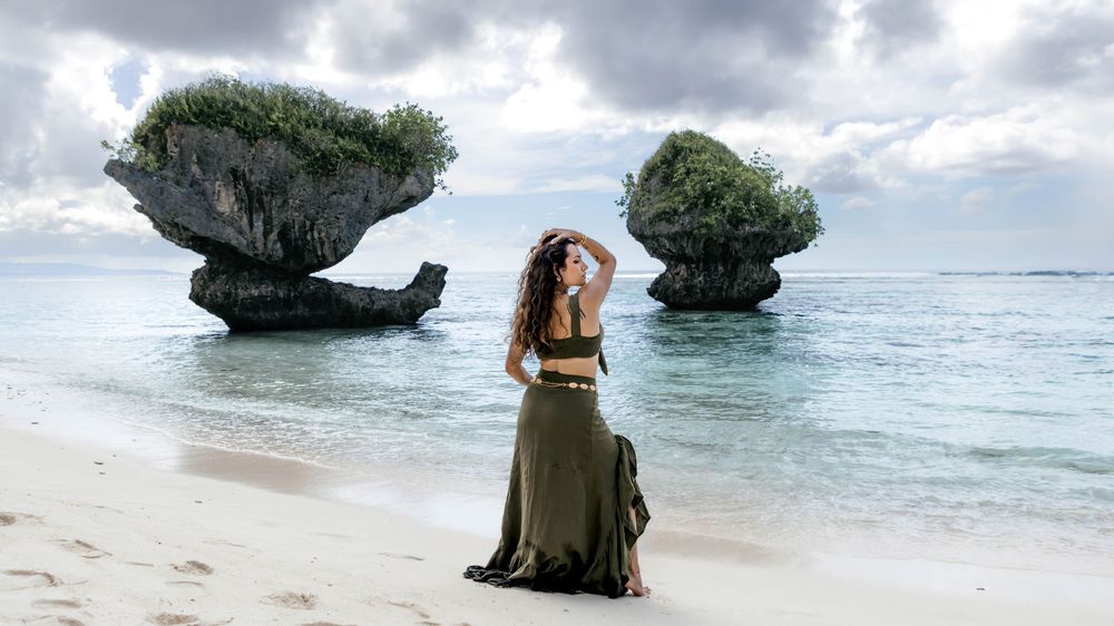 Woman in olive maxi skirt poses on white sand beach with dramatic mushroom-shaped rock formations rising from turquoise sea.