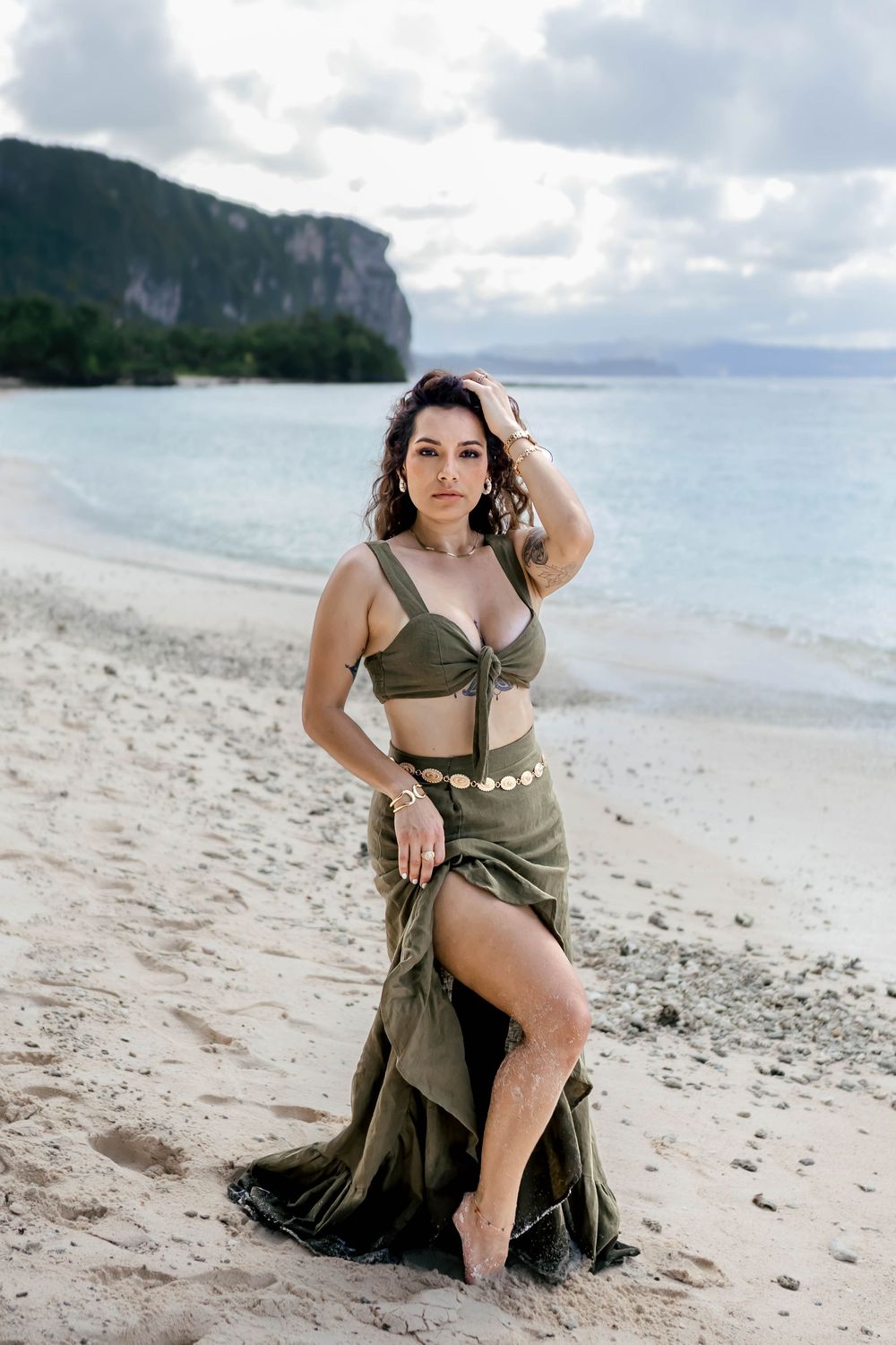 Woman in olive green two-piece outfit posing on sandy beach with turquoise water and rocky cliffs behind her.