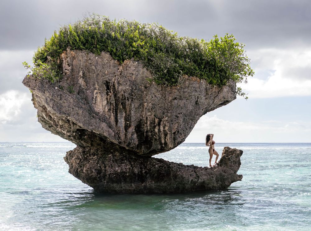 Woman standing on a dramatic mushroom-shaped rock formation rising from turquoise ocean waters, topped with lush green tropical vegetation.