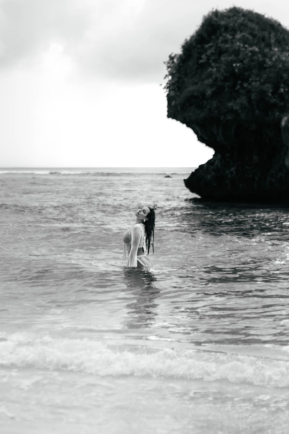 Black and white photo of a person wading in ocean waves near a rocky outcrop on a tropical beach.