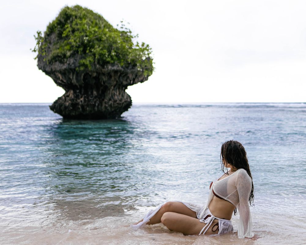 Woman in bikini sitting on a tropical beach shore gazing at a mushroom-shaped rock formation rising from turquoise waters.