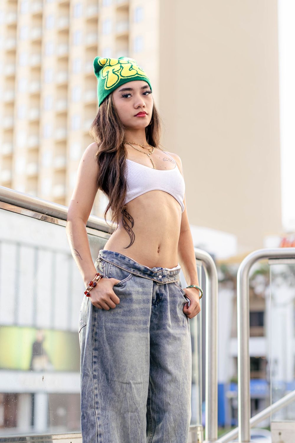 Young woman in white crop top, baggy jeans, and green beanie poses outdoors near metal railing with urban backdrop.