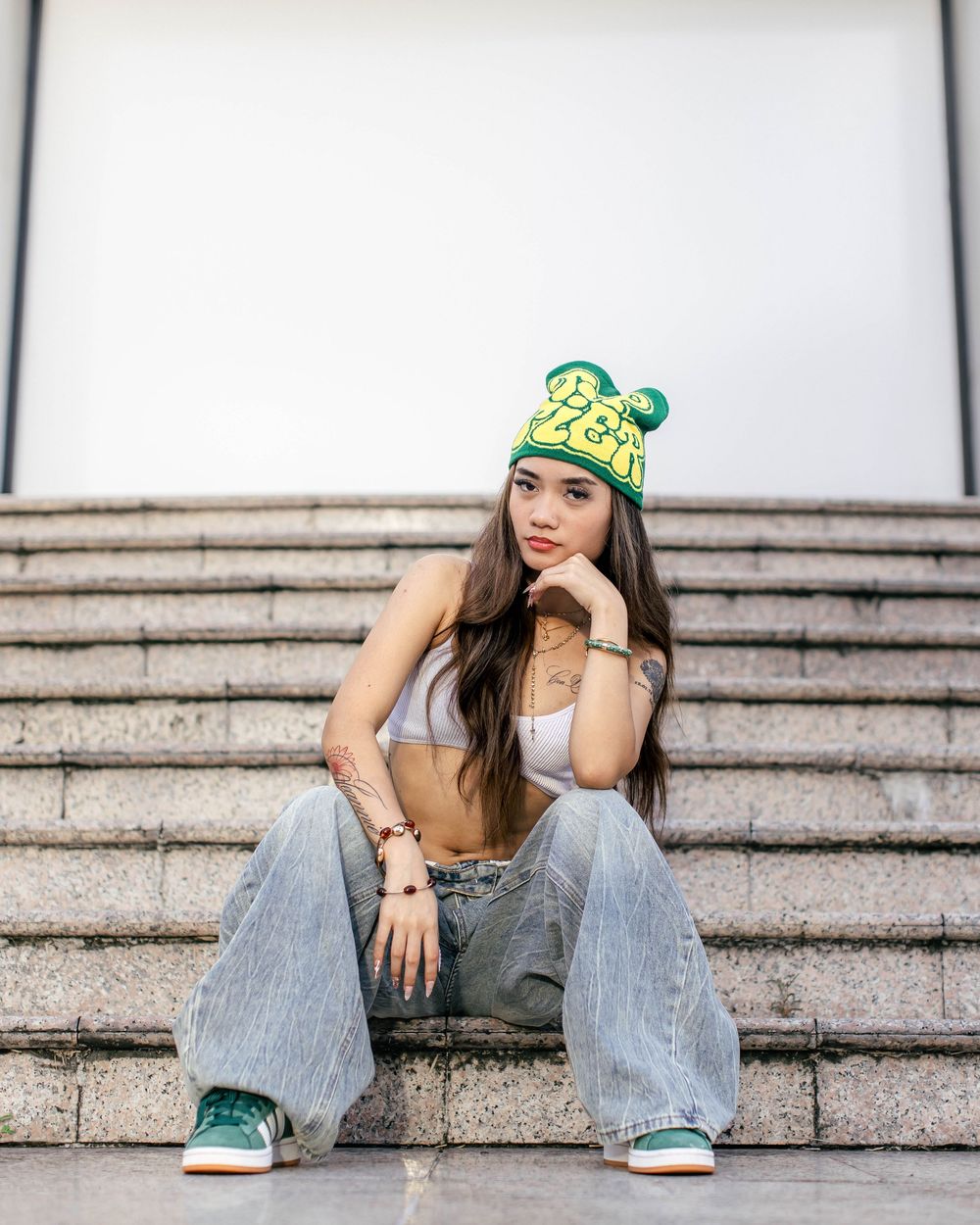 Young woman in green beanie, crop top, wide-leg jeans, and green sneakers sitting on outdoor steps.