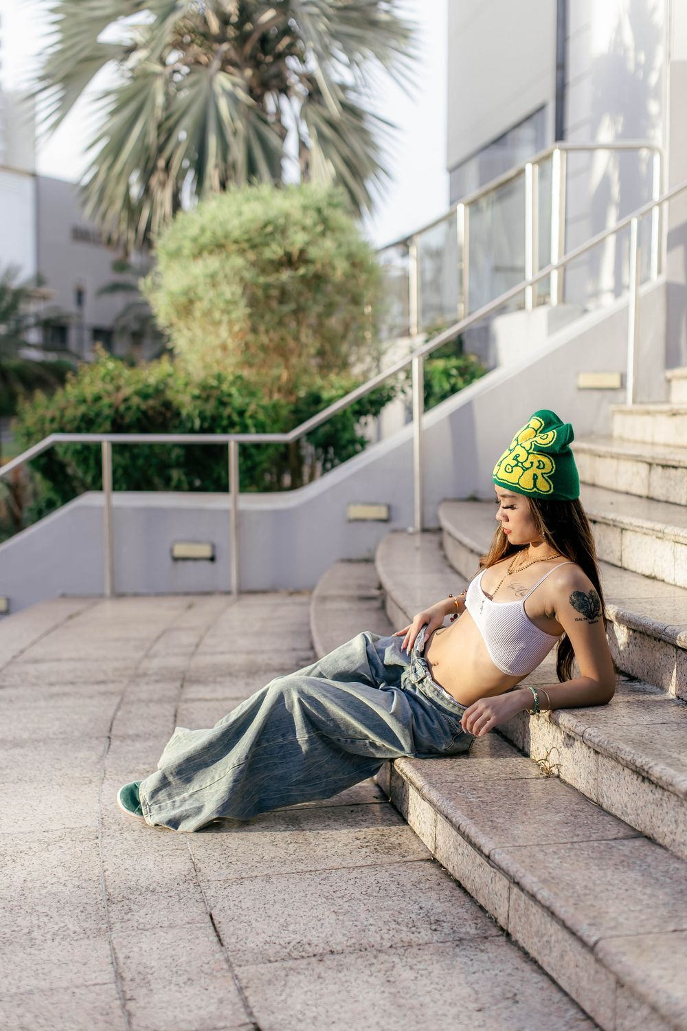 Young woman in white crop top, baggy jeans, and green beanie sits on outdoor steps near palm trees.