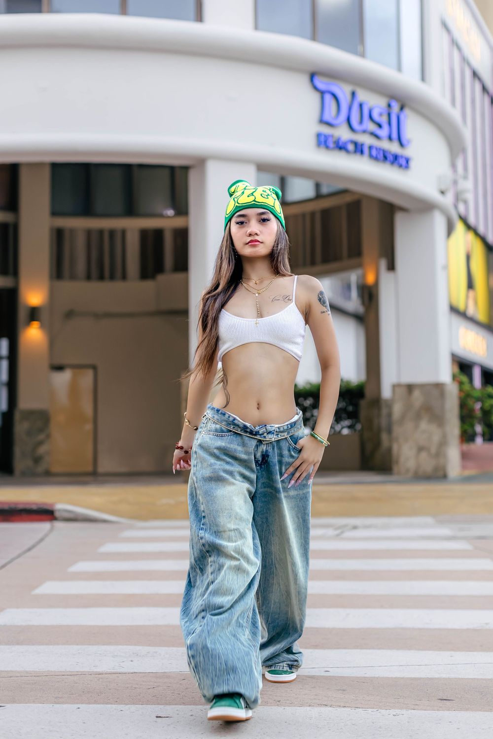 Young woman in white crop top, baggy jeans, and green beanie walks confidently across a crosswalk near Dusit Beach Resort.