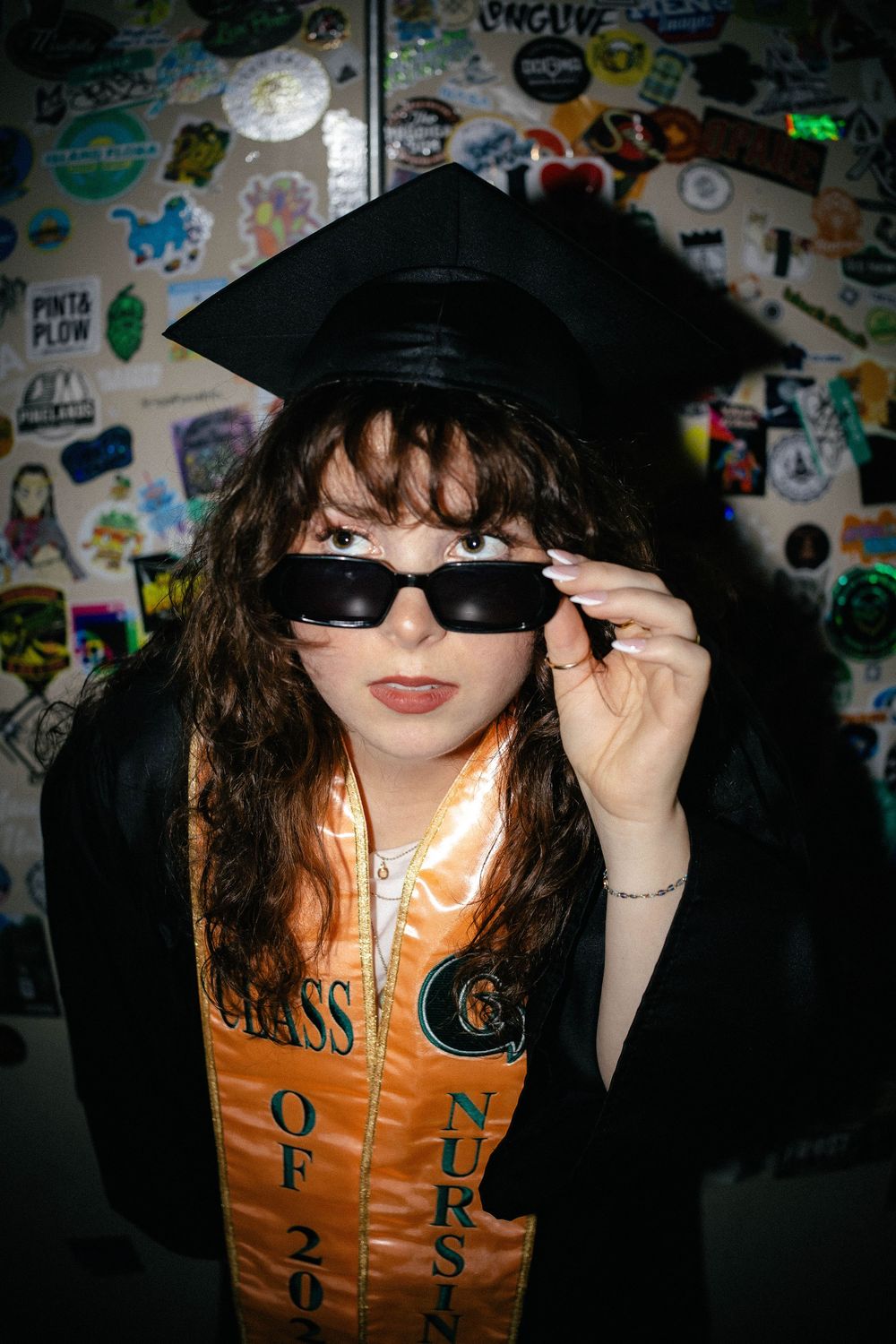 Graduate in black cap and gown with orange nursing sash poses coolly with sunglasses against sticker-covered wall.
