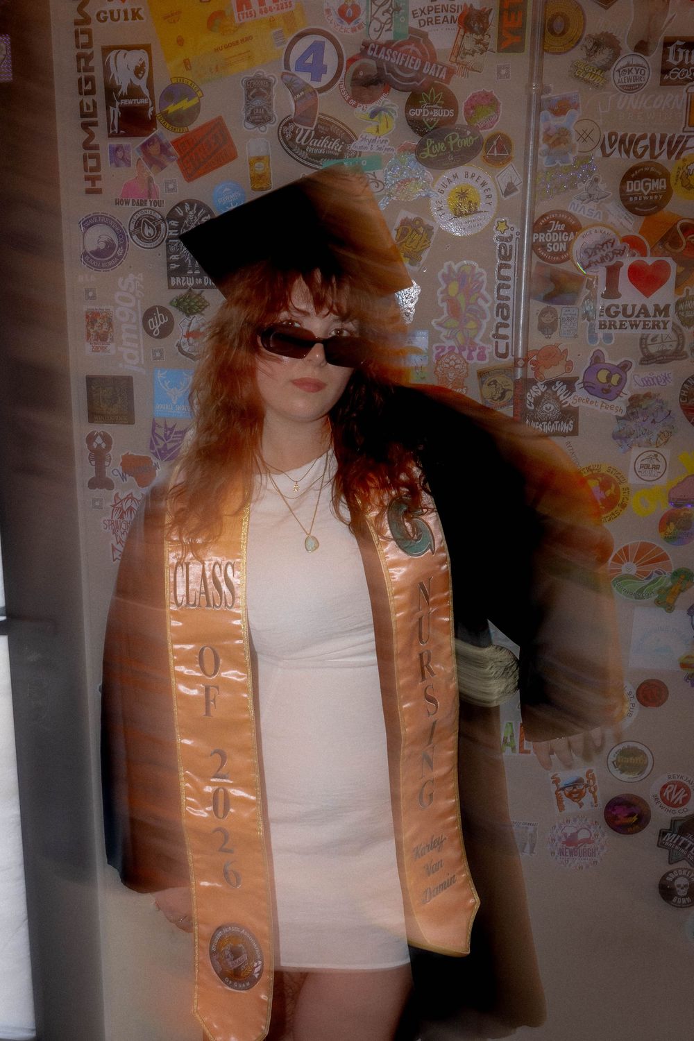 A young woman in graduation cap and gold CLASS OF 2024 sash poses in white dress and sunglasses before a sticker-covered wall.