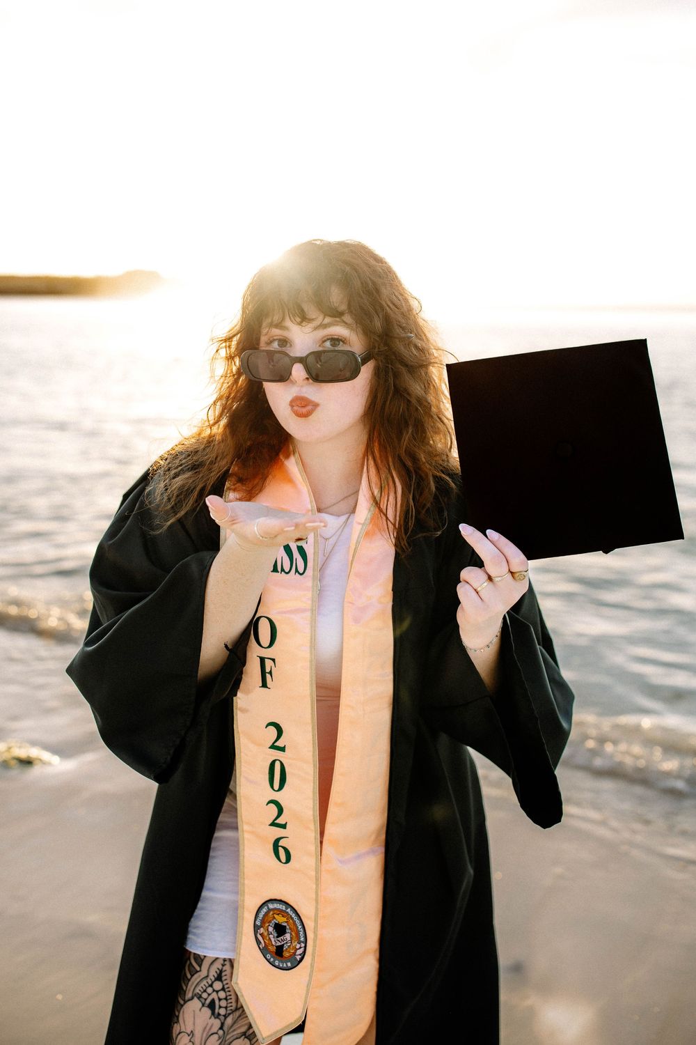 Graduate in black gown and Class of 2026 sash holds diploma at beach sunset, blowing a kiss wearing sunglasses.