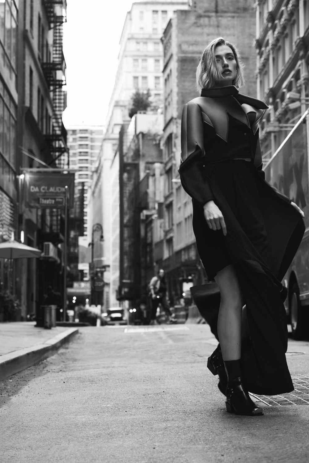 Dramatic black and white fashion photograph of a flowing black dress on a moody city street with tall buildings.