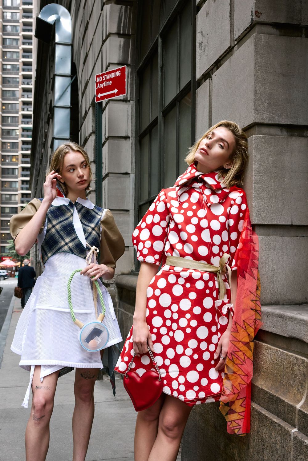 Two figures in contrasting outfits pose against stone building wall, one wearing polka dot dress and one in plaid with apron.