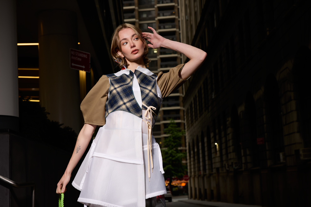 Young woman in plaid top and white skirt poses outside modern building at night.