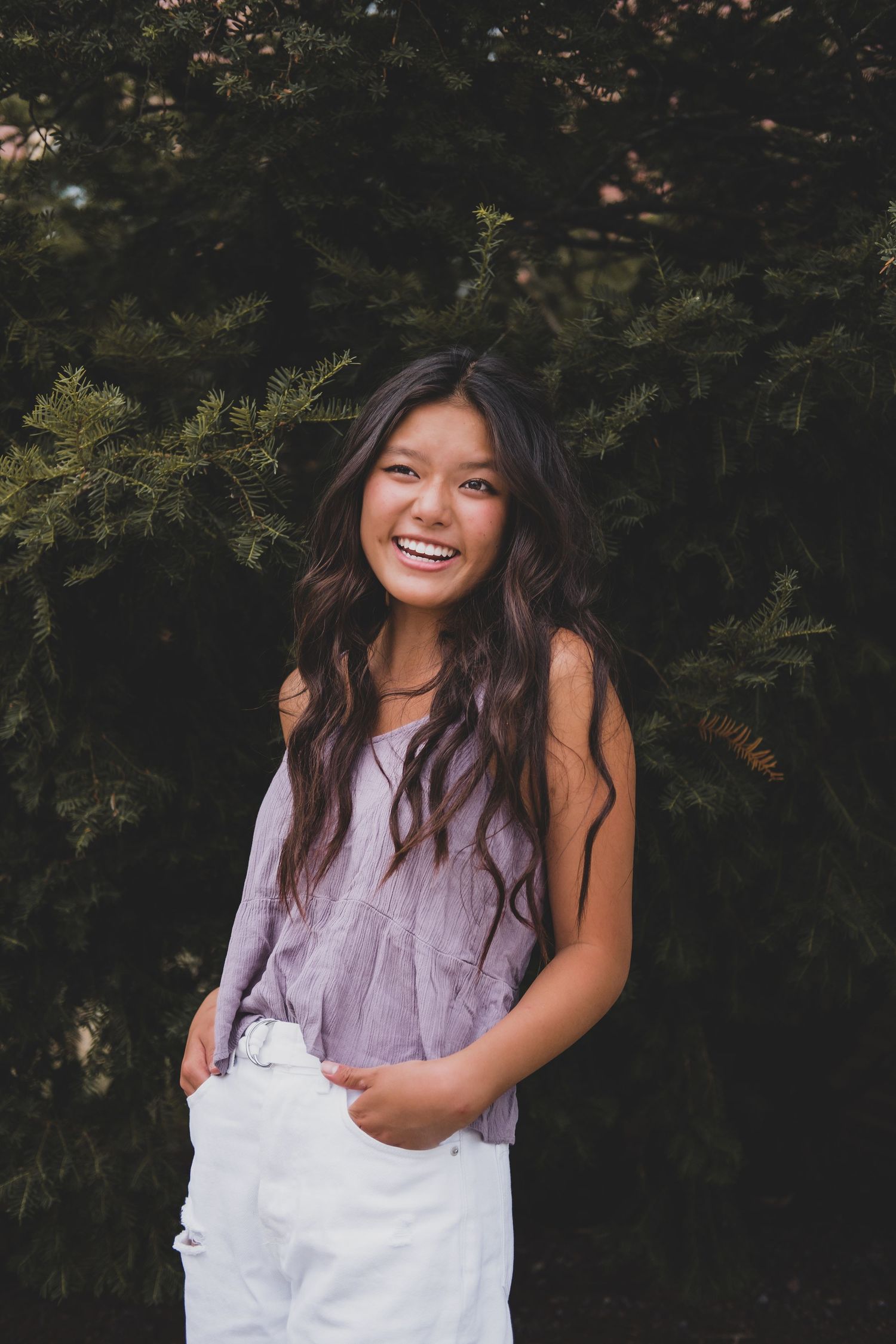 A person in a lavender top and white overalls poses playfully against a dark background.