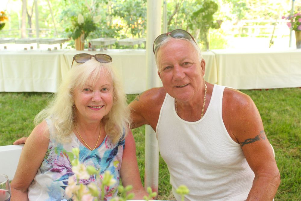 A couple smiles at outdoor event wearing summer attire under white tent.