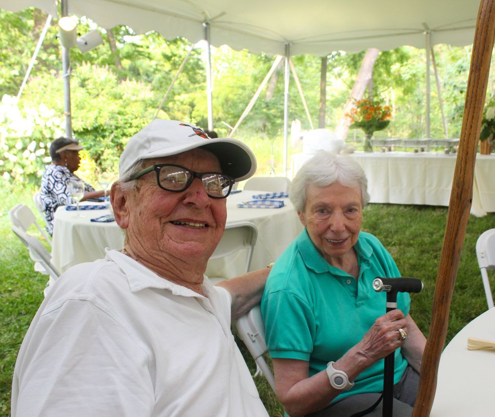 Two seniors seated under white tent at outdoor gathering.