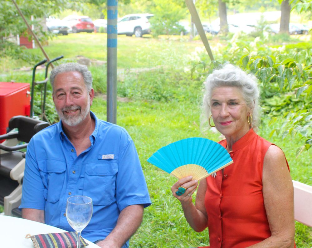 A couple enjoying an outdoor gathering, with woman holding blue fan in coral top.