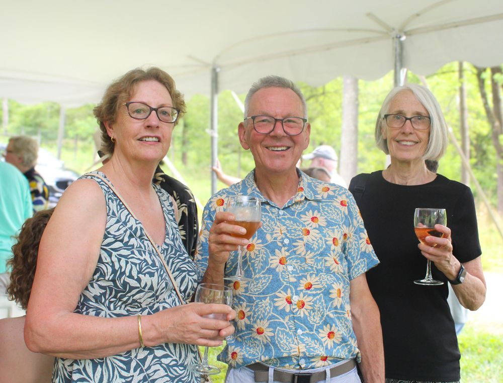 Three people enjoying drinks and conversation under a white tent at an outdoor social gathering.
