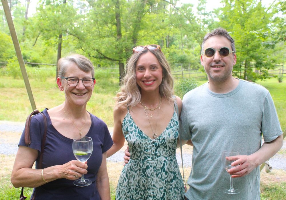 Three people posing with drinks at a casual outdoor gathering surrounded by green trees.