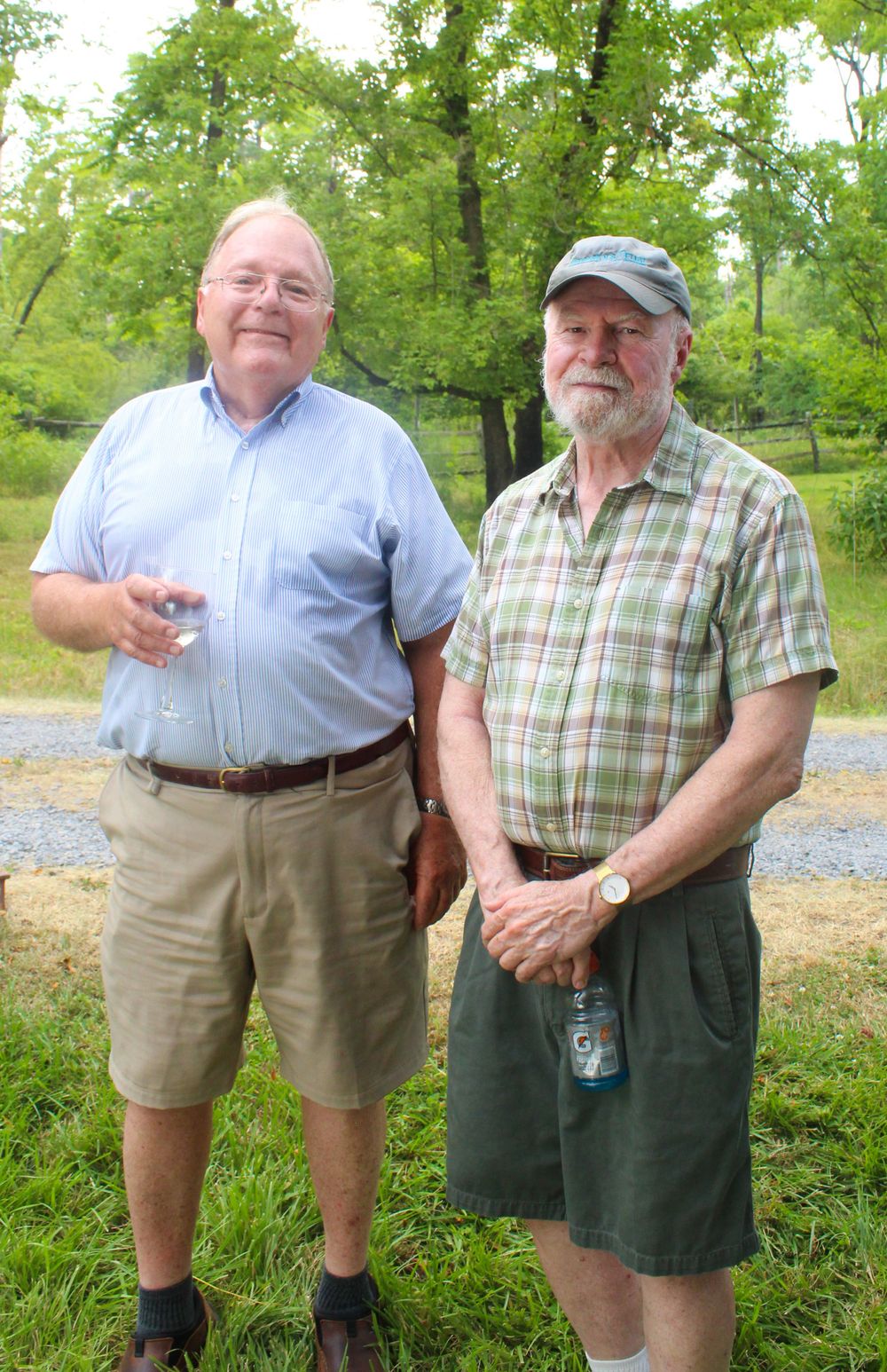 Two men in casual summer attire standing together on a gravel path with trees in background.