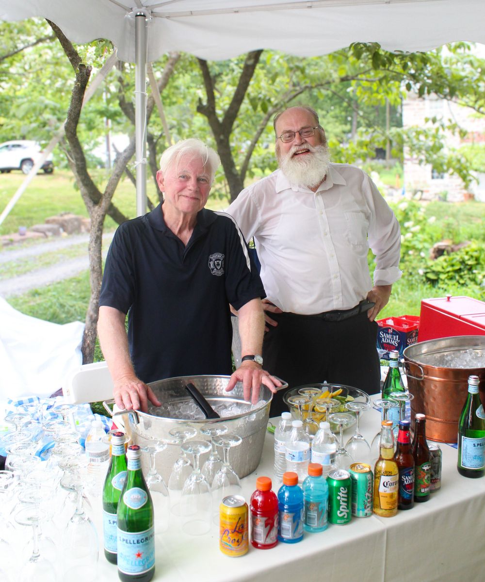 Two men staffing a beverage station with assorted drinks and ice bucket under a white tent.