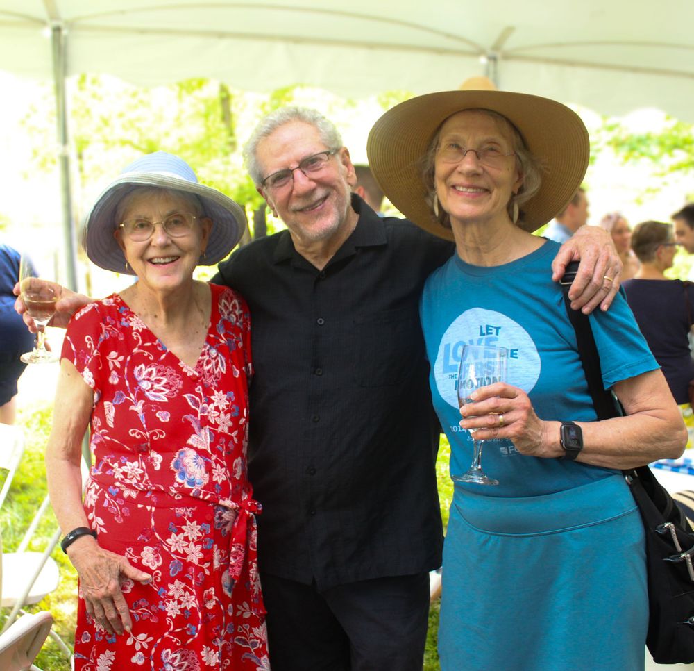 Three people wearing summer hats smiling together under a white tent at an outdoor gathering.