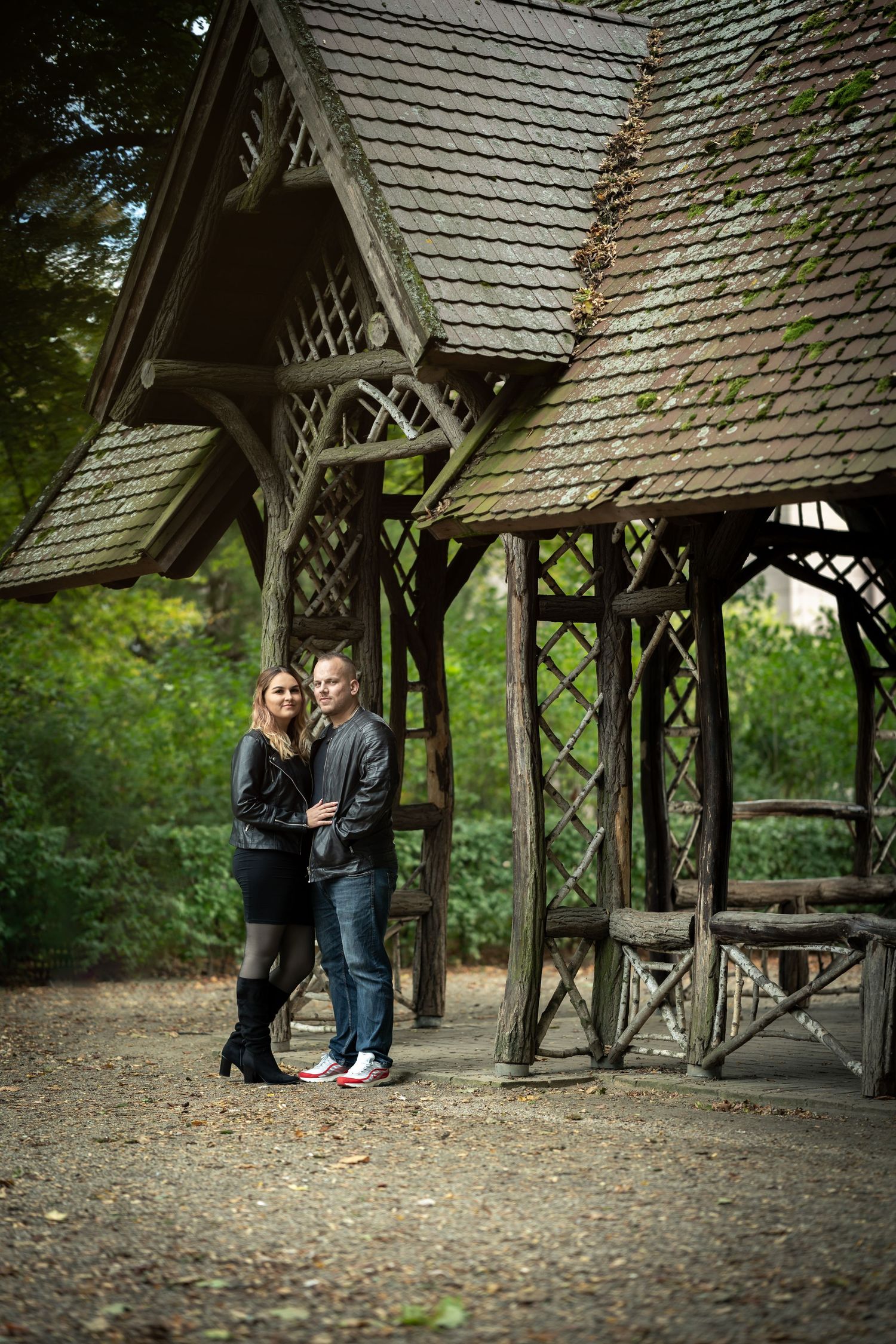 A rustic wooden gazebo with shingled roof stands in a peaceful wooded setting on a dirt path surrounded by greenery.