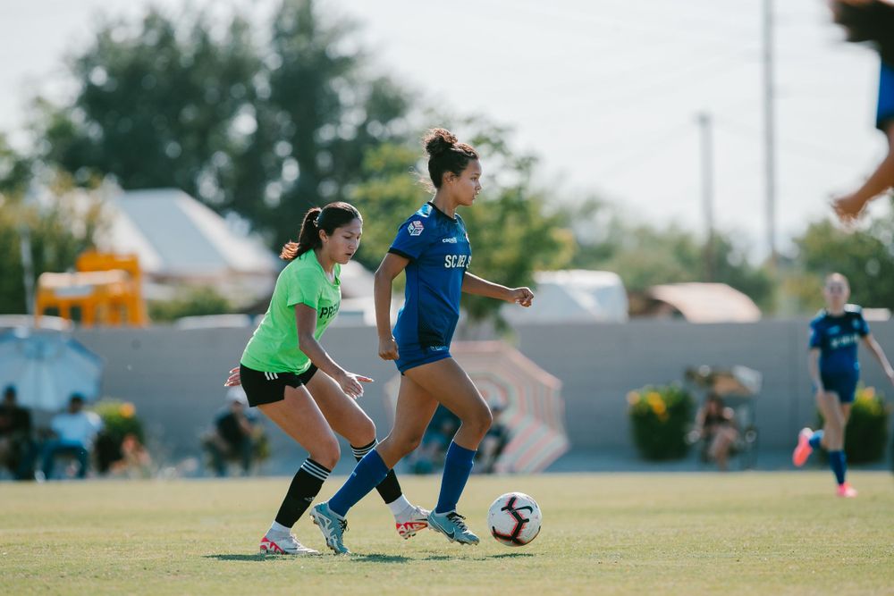 Two female soccer players competing for ball during outdoor match in blue and green uniforms.