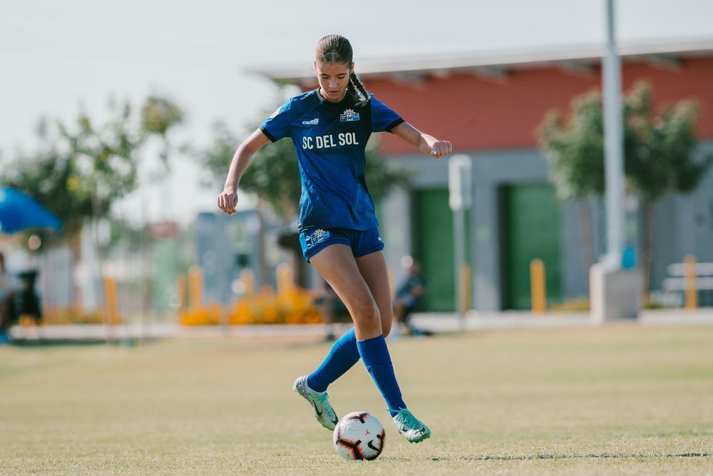 Female soccer player in blue FC Del Sol uniform dribbling ball on sunny outdoor field.