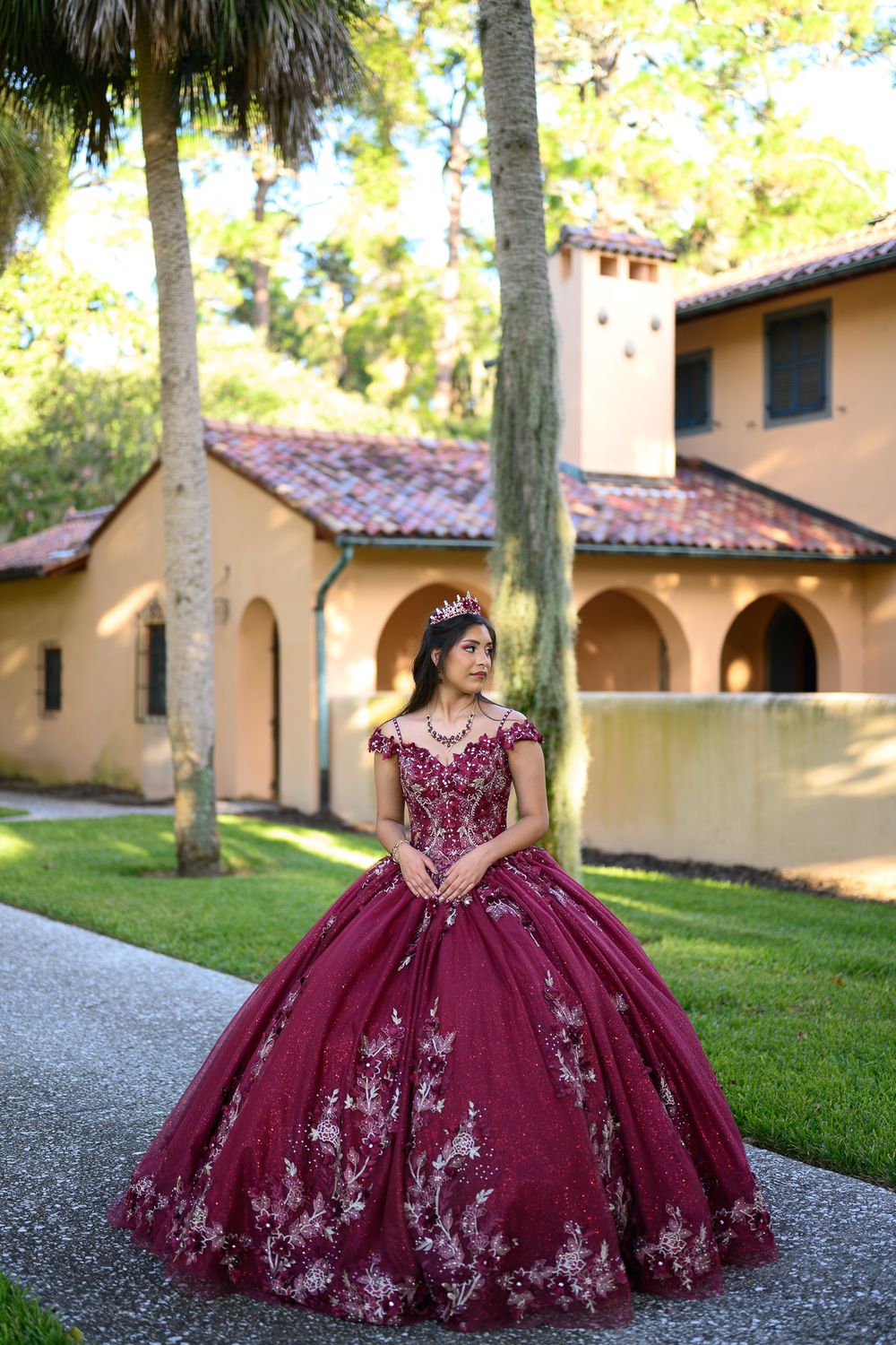 Young woman in elegant burgundy quinceañera ball gown with floral embroidery poses outdoors near Spanish-style building.