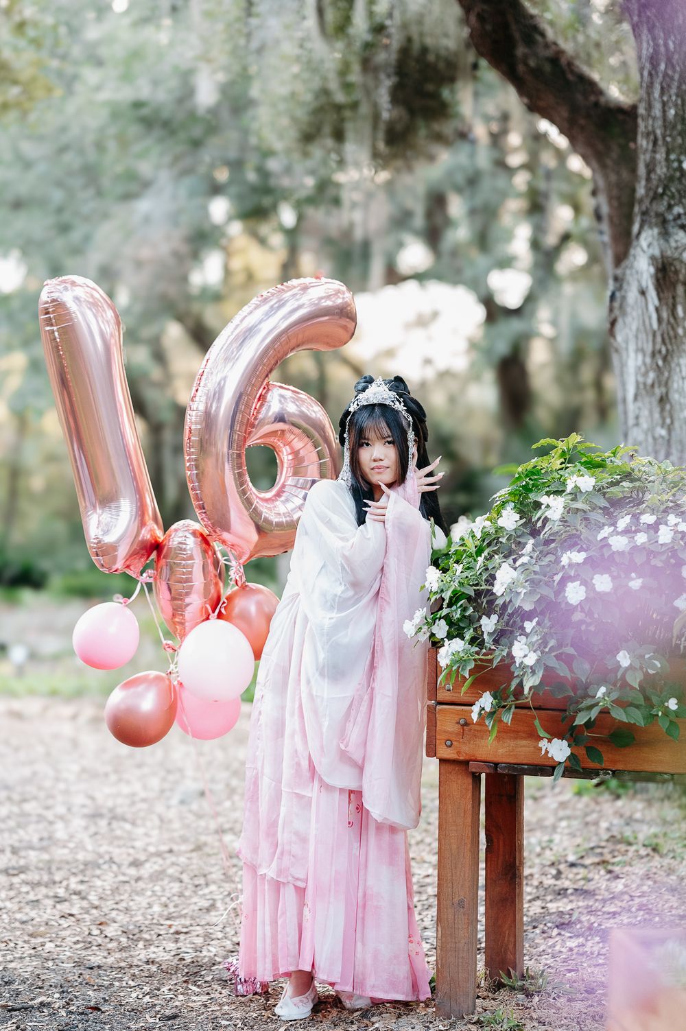 Someone in a pink traditional hanfu dress poses with rose gold number balloons and floral decorations in a dreamy outdoor setting.