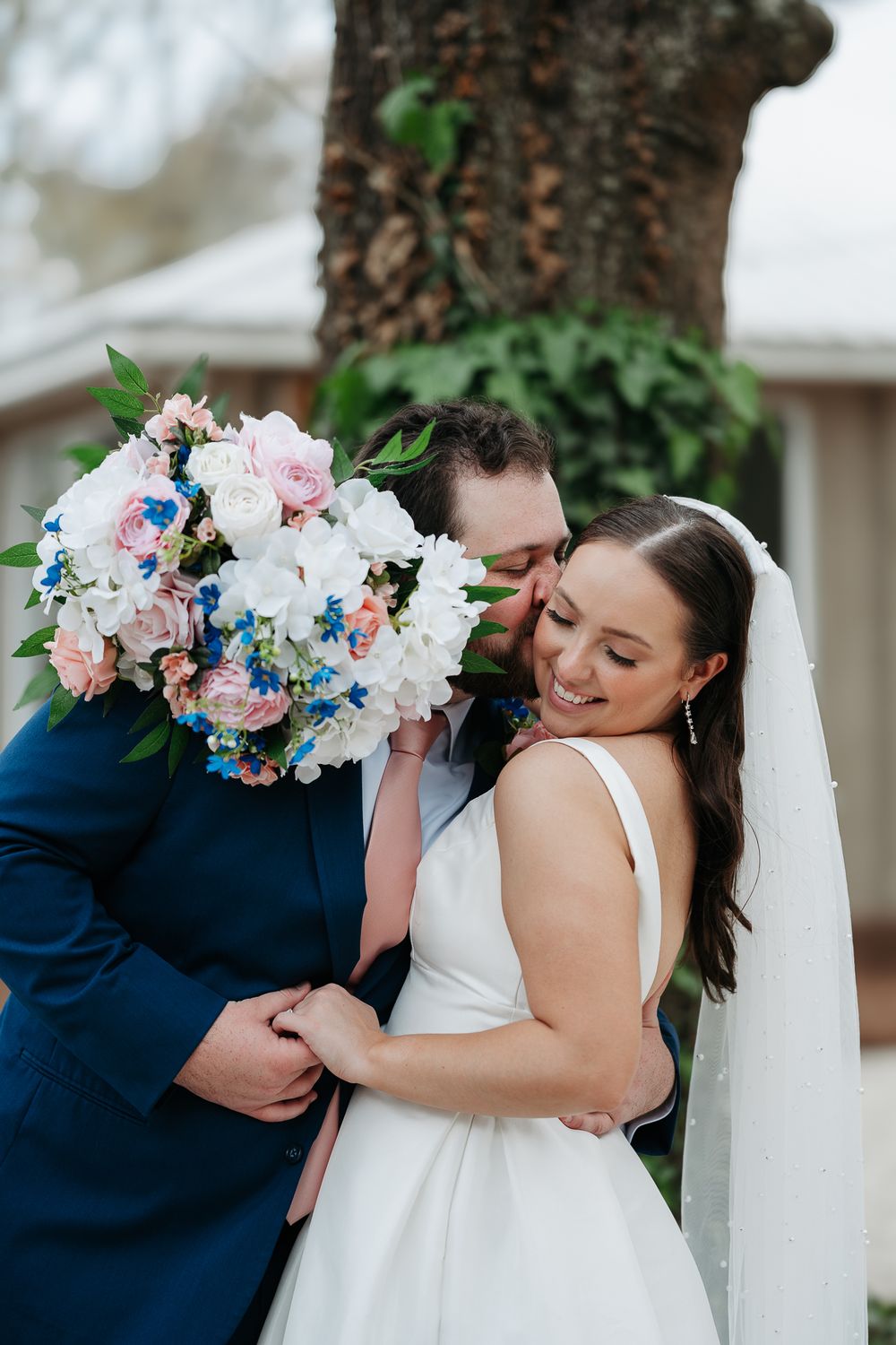 Happy wedding moment captured outdoors with beautiful bouquet featuring white and pink peonies with blue accents.