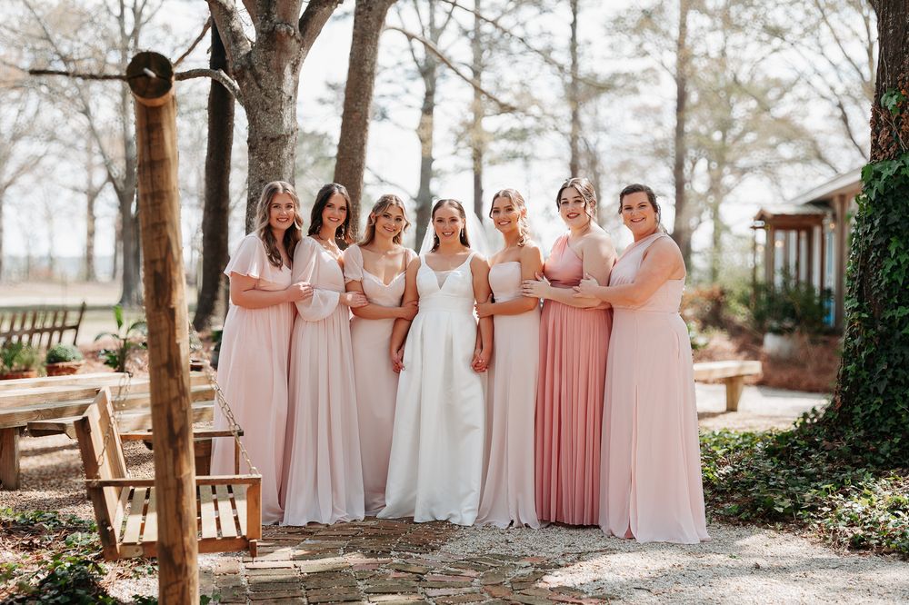 A wedding party poses outdoors in blush pink and white dresses holding bouquets against a wooded backdrop.