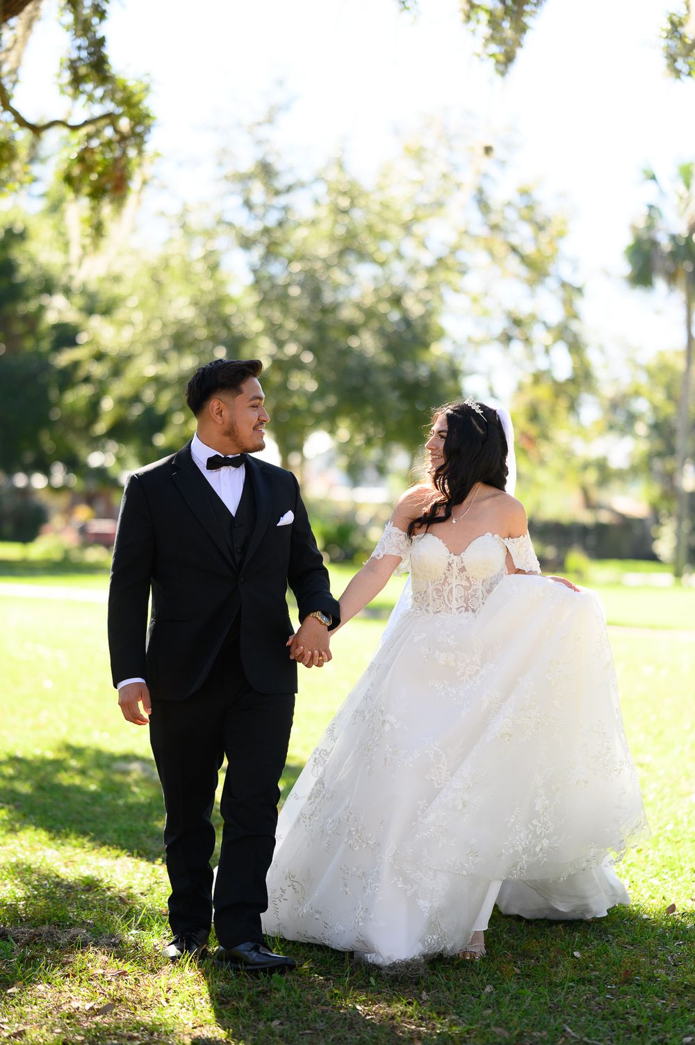 Newlywed couple walks hand in hand through a sunlit garden in formal wedding attire.