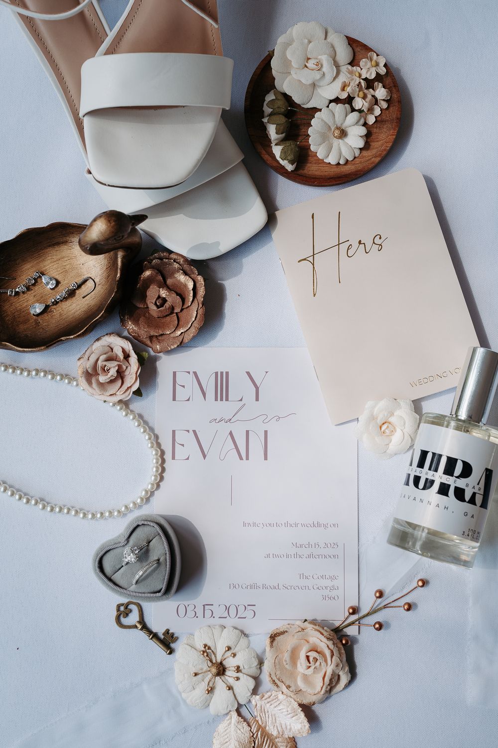 Elegant wedding details flatlay with white shoes, invitation, perfume bottle, pearl necklace, key and floral decorations.
