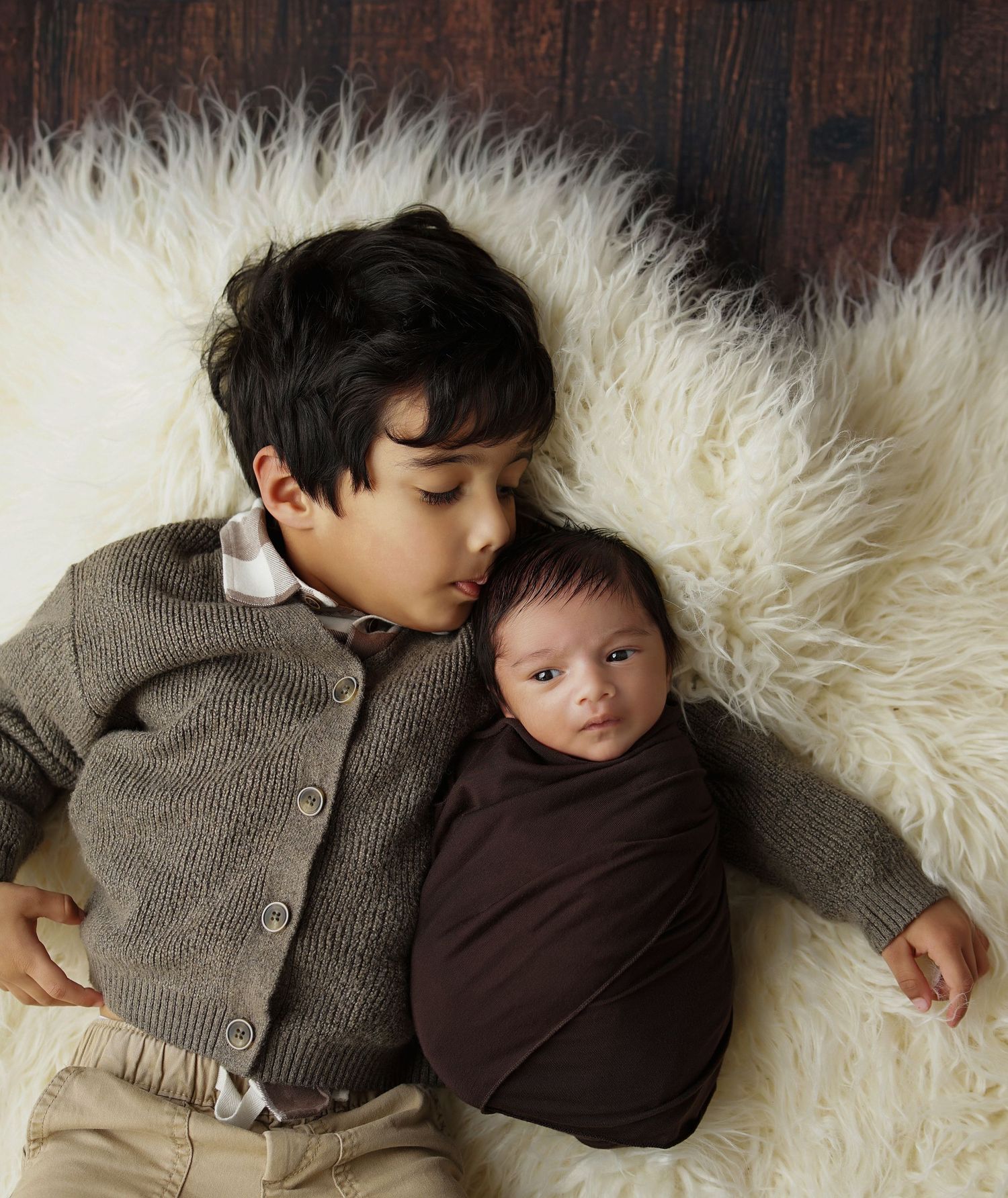 Young boy in a gray cardigan gently kisses his swaddled newborn sibling on a soft white fur rug in a sweet sibling newborn portrait.