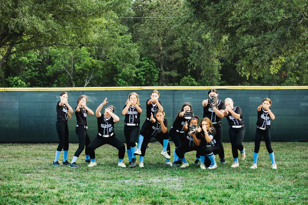 Girls softball team in black uniforms posing together on a green field with trees in the background.